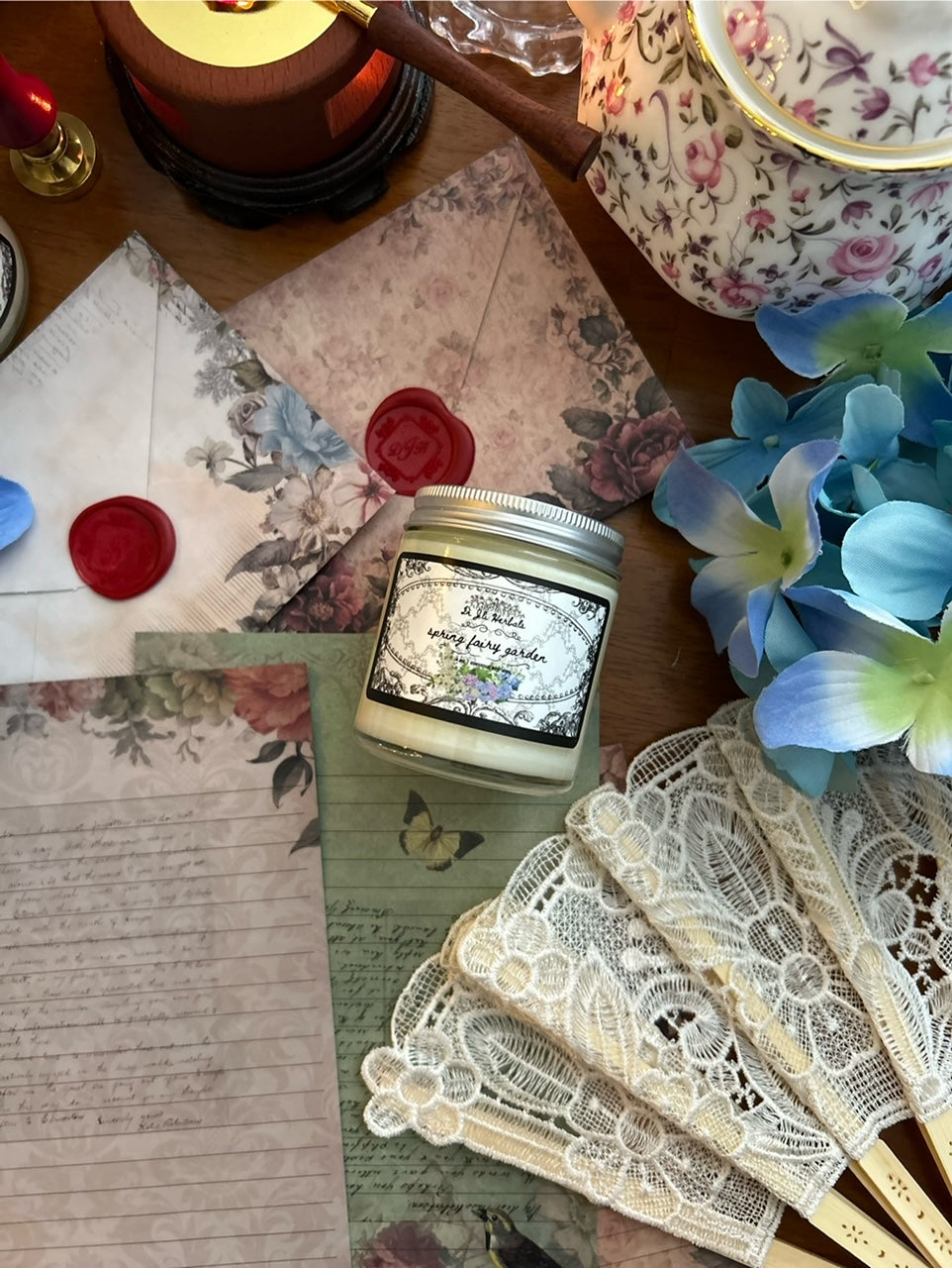 Lit floral scented storybook candle in a victorian romantic-inspired jar surrounded by a white lace fan, flowers, botanically decorated stationery and red wax seals, alongside Wedgewood china tea cup on a wooden table. 