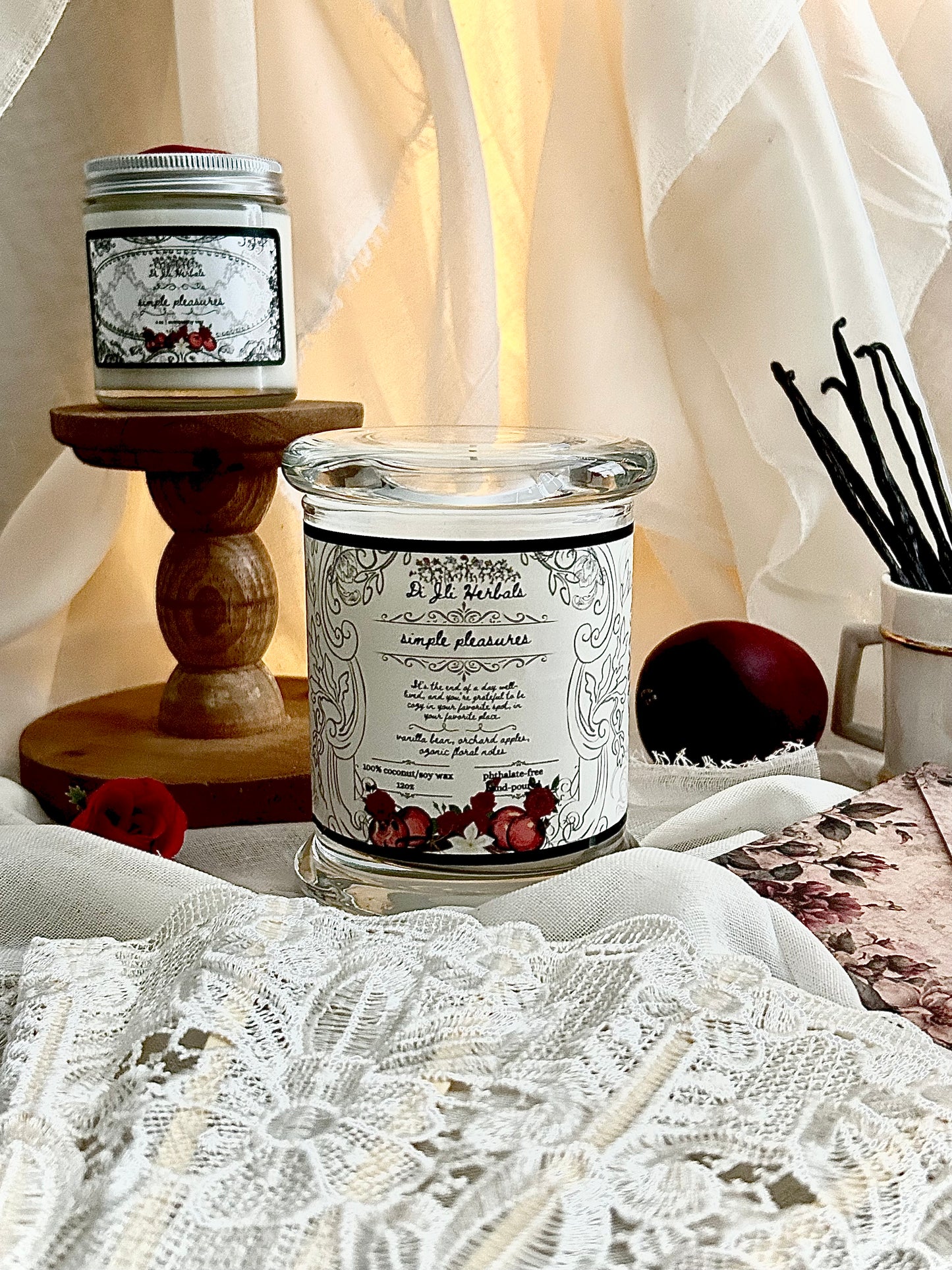 Hand-poured Victorian romantic-inspired candle with elegantly designed simple pleasures label on a white lace tablecloth, an apple and a cup of vanilla beans in the background.