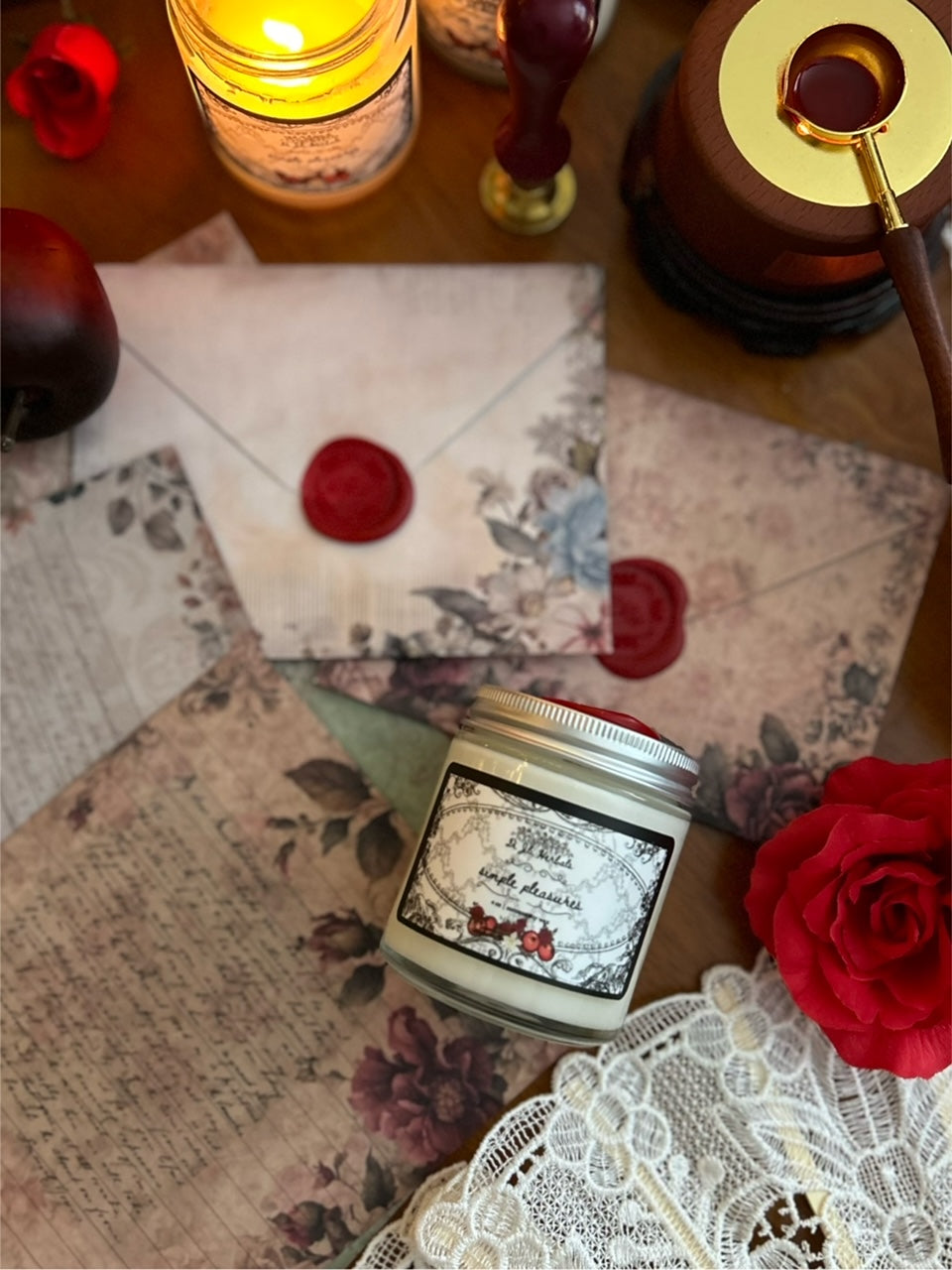 Lit apple gourmand scented candle in a victorian romantic-inspired jar surrounded by a white lace fan, apples, roses, botanically decorated stationery, red wax seals, and a Wedgewood china tea cup on a wooden table. 