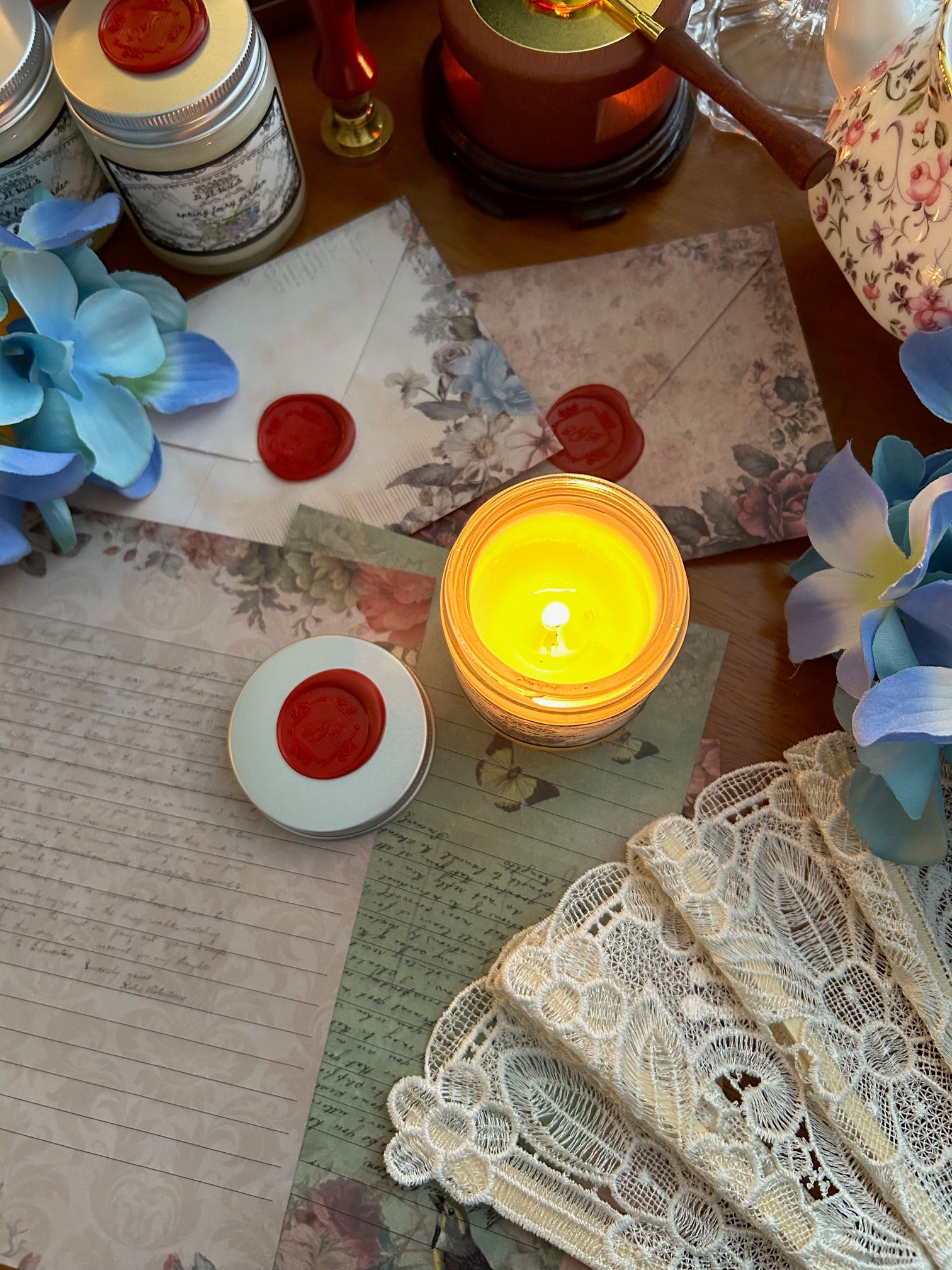 Lit spring floral scented candle in a victorian romantic-inspired jar surrounded by a white lace fan, flowers, botanically decorated stationery and red wax seals, alongside Wedgewood china tea cup on a wooden table. 