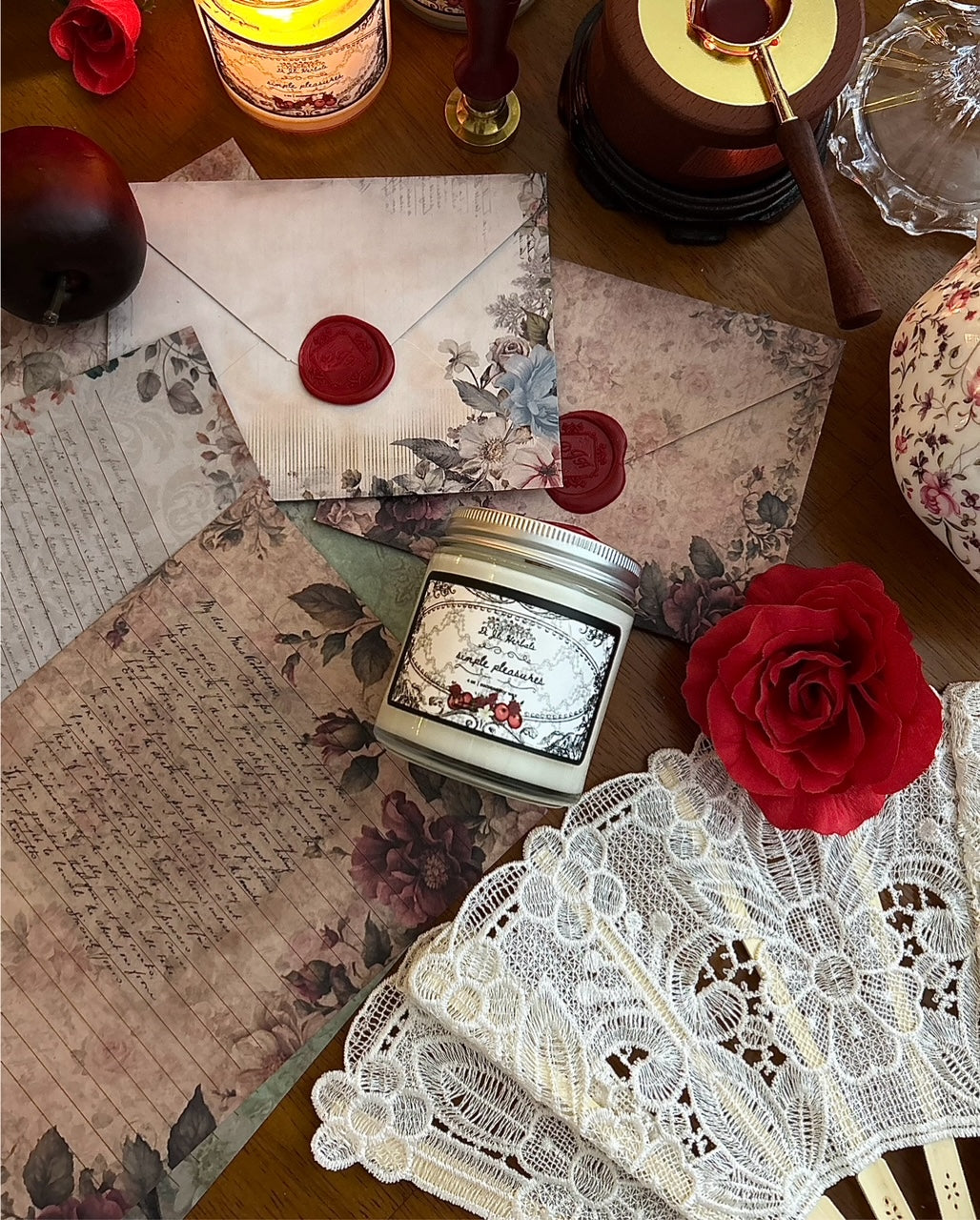 Lit apple gourmand scented candle in a victorian romantic-inspired jar surrounded by a white lace fan, apples, roses, botanically decorated stationery and red wax seals, alongside Wedgewood china tea cup on a wooden table. 