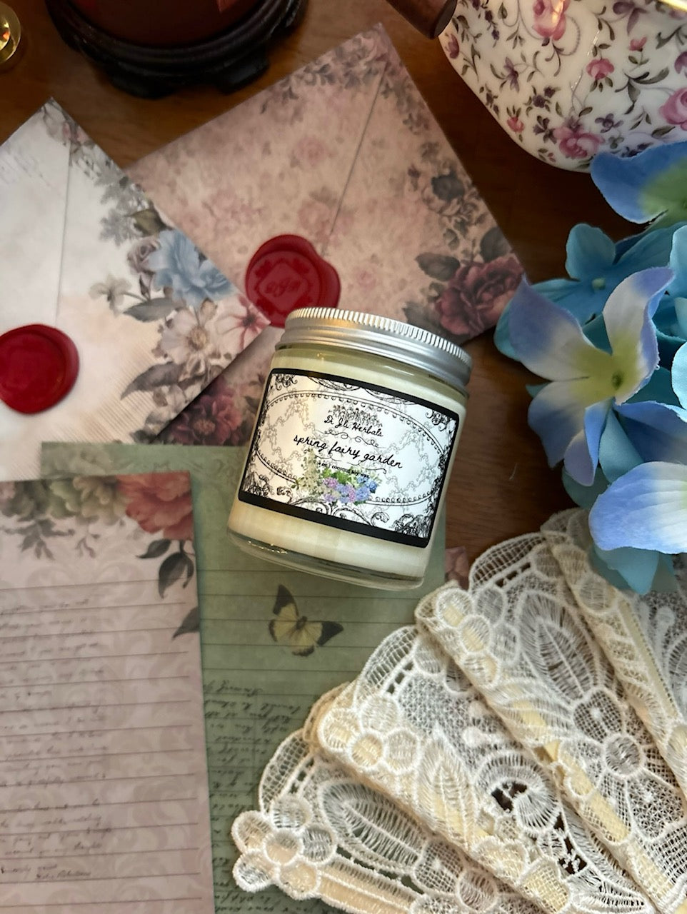 Lit spring garden scented candle in a victorian romantic-inspired jar surrounded by a white lace fan, pinecones, botanically decorated stationery and red wax seals, alongside Wedgewood china tea cup and flowers on a wooden table. 